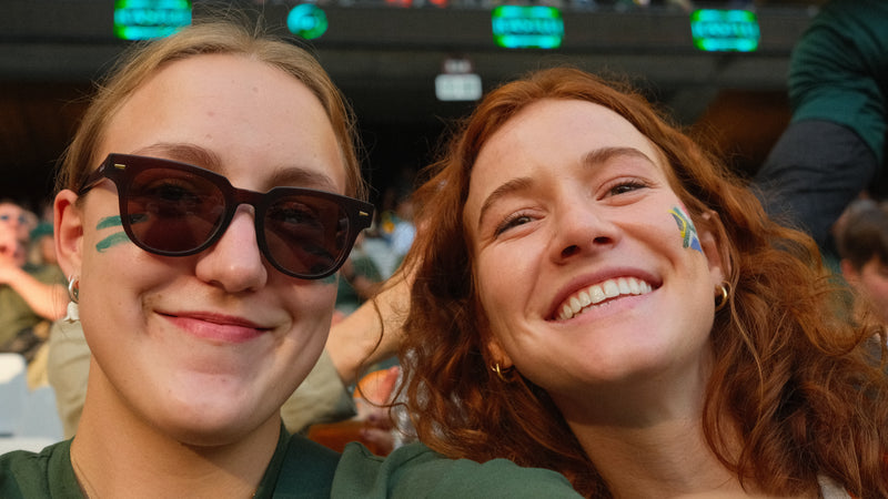 Two women with sunglasses and painted faces in a stadium setting