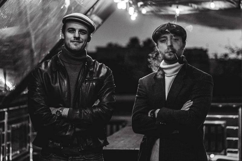 Two men standing side by side in a black and white photo, with one wearing a leather jacket and the other in a suit.