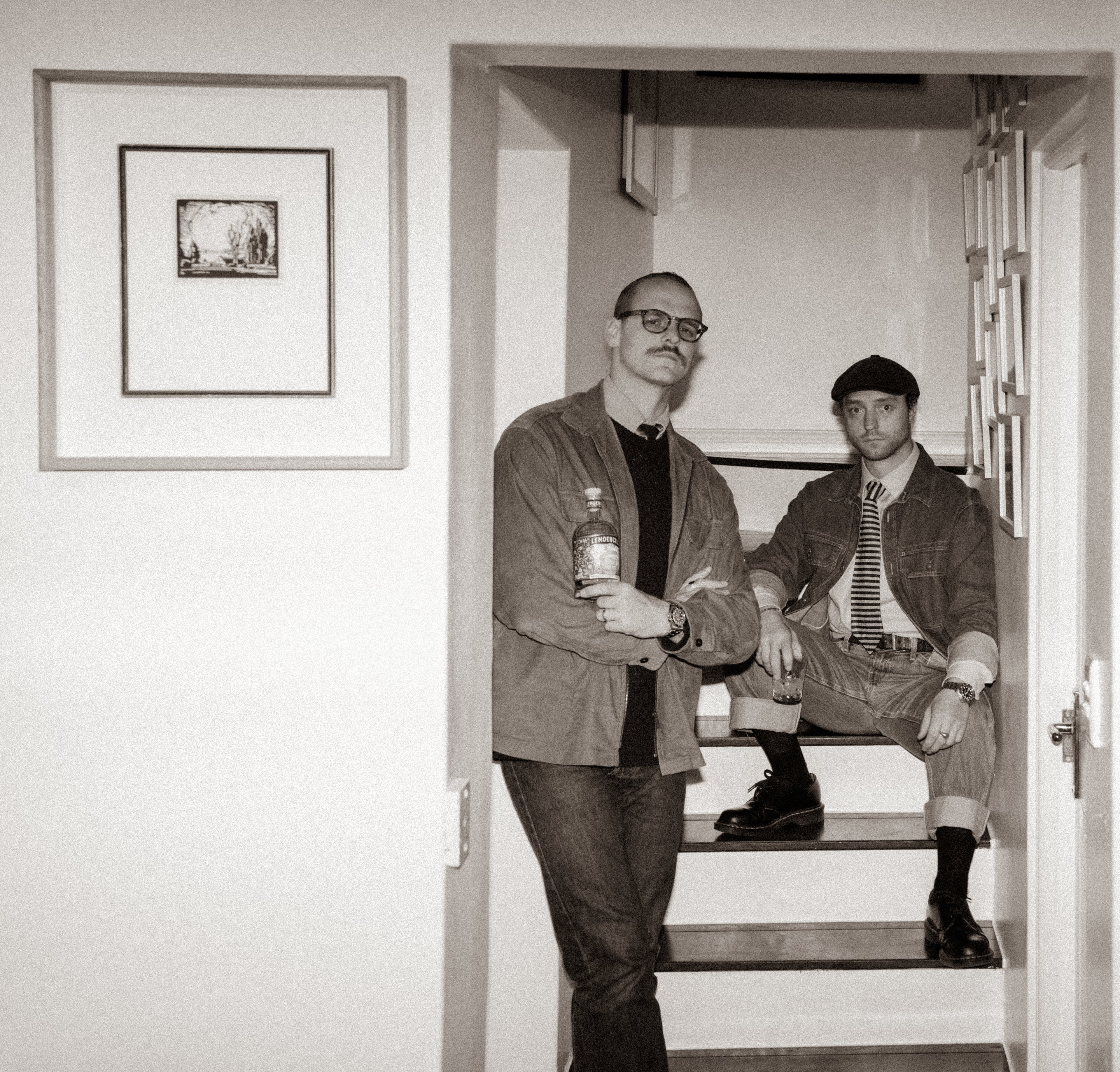 Danie en Moskou du Toit from Spoegwolf standing on a staircase holding a bottle of Lemoencello, with a framed Pierneef on the wall.