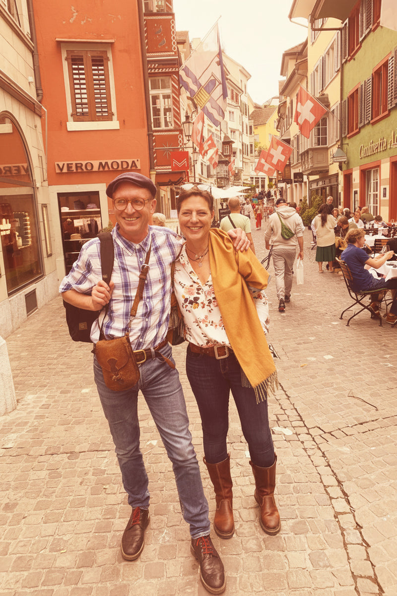 Two people posing on a street with colorful buildings and flags in the background