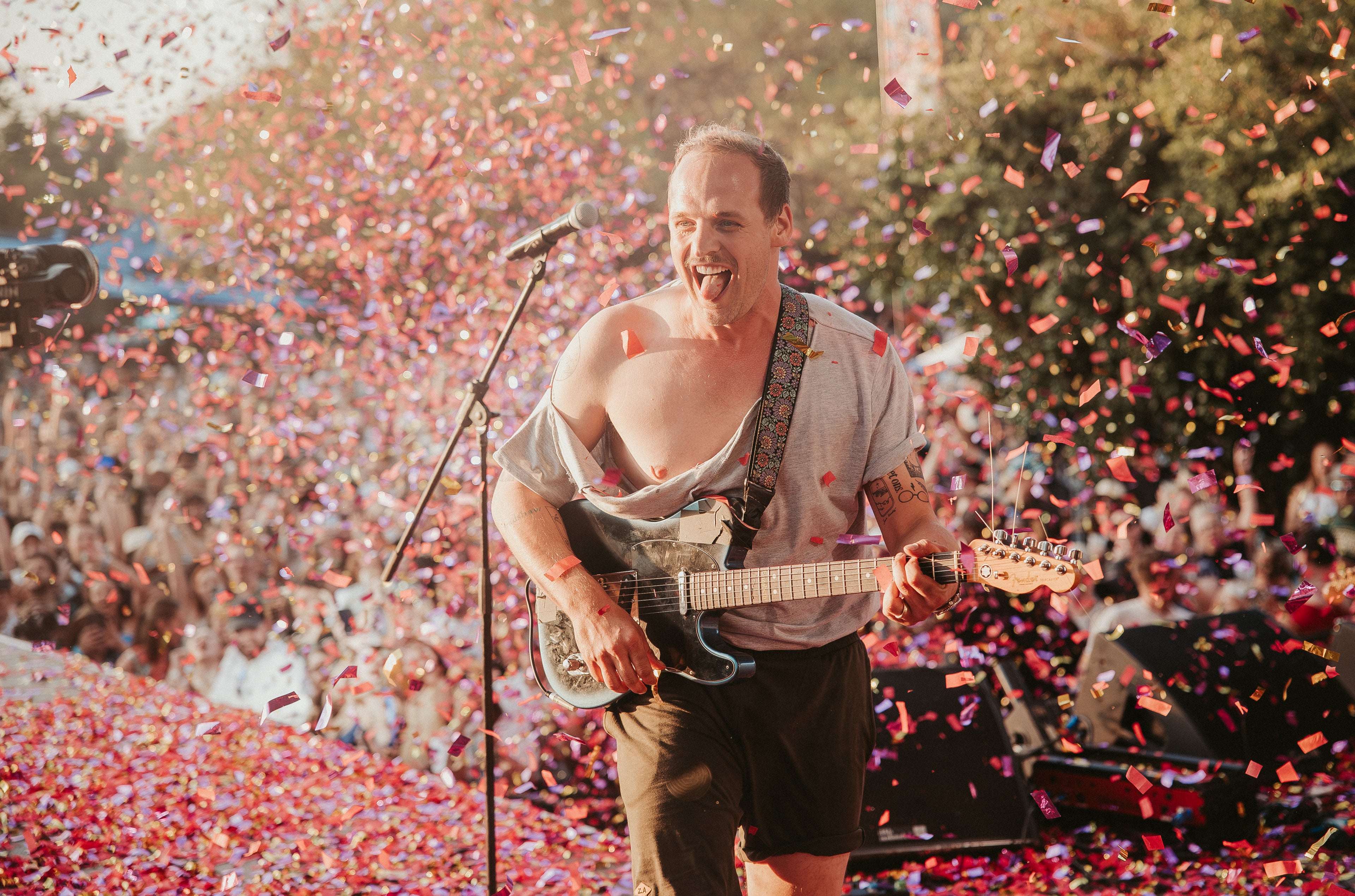 Man playing guitar on stage with confetti falling, surrounded by a crowd.