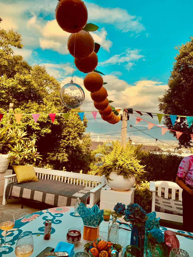 Outdoor setting with a table set for a meal, balloons, oranges, and a scenic background.