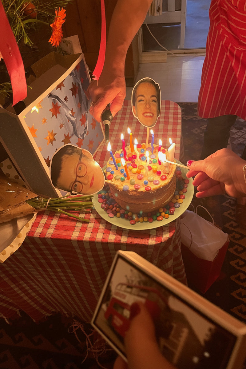Birthday cake with lit candles on a checkered tablecloth, with people holding cutout faces.