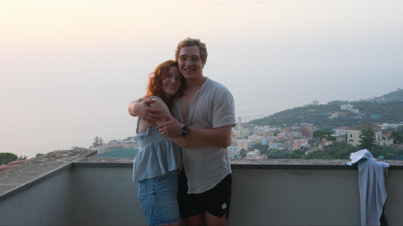 Two people standing on a rooftop in Italy with a scenic view of a town and mountains in the background.
