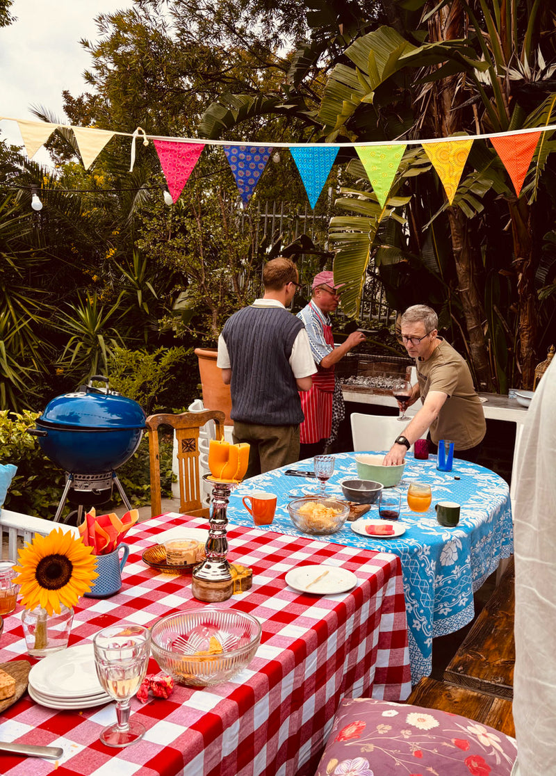 Outdoor gathering with people around a table set for a meal, decorated with colorful flags and a barbecue grill.