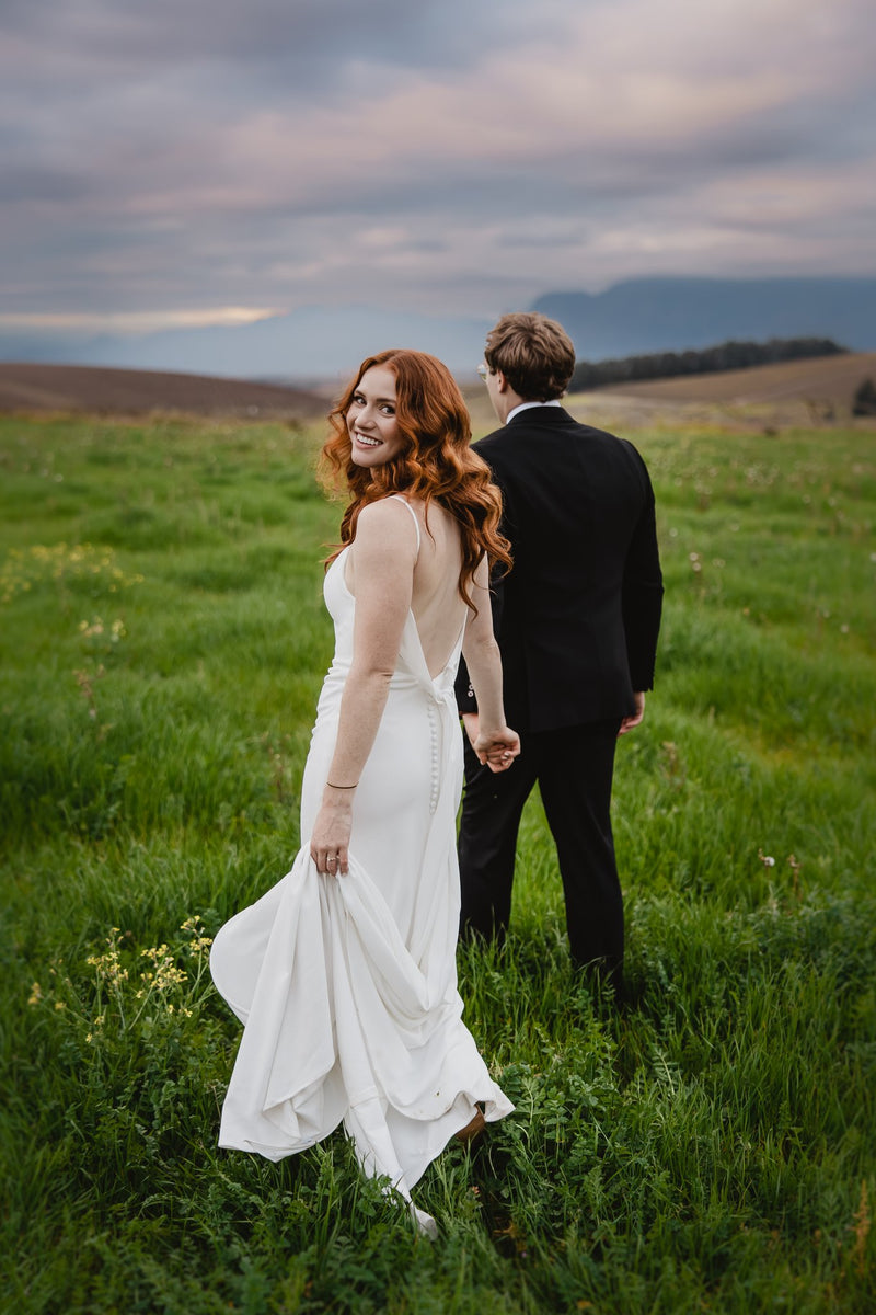 Couple in wedding attire standing in a grassy field with mountains in the background
