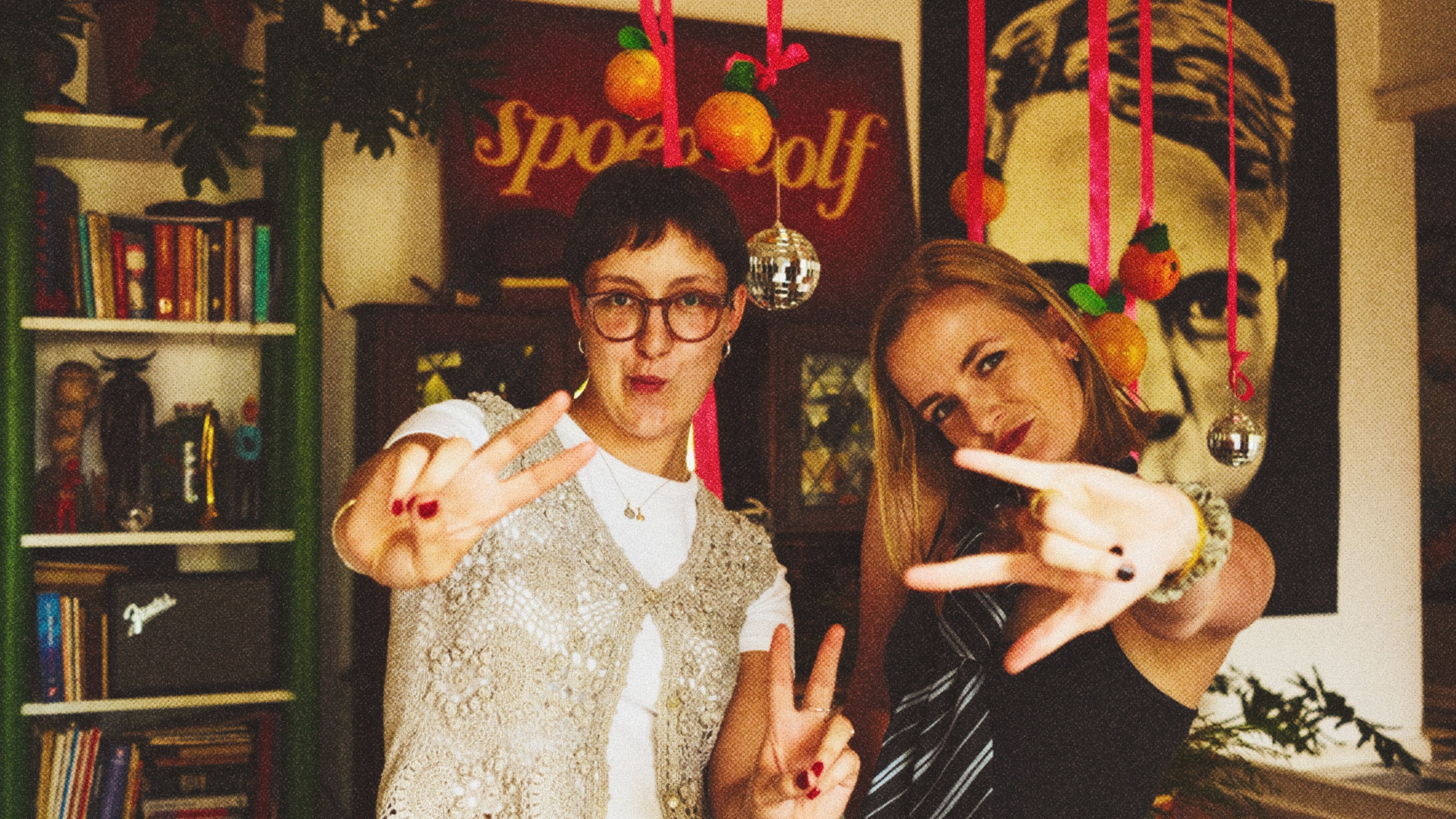 Two women making peace signs in a room with bookshelves and a 'Spoegwolf' sign, and oranges hanging in the air.