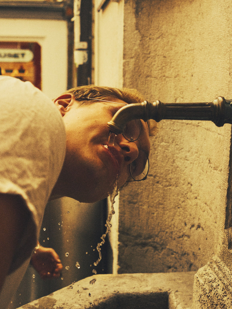 Person drinking water from an outdoor faucet with a stone wall background